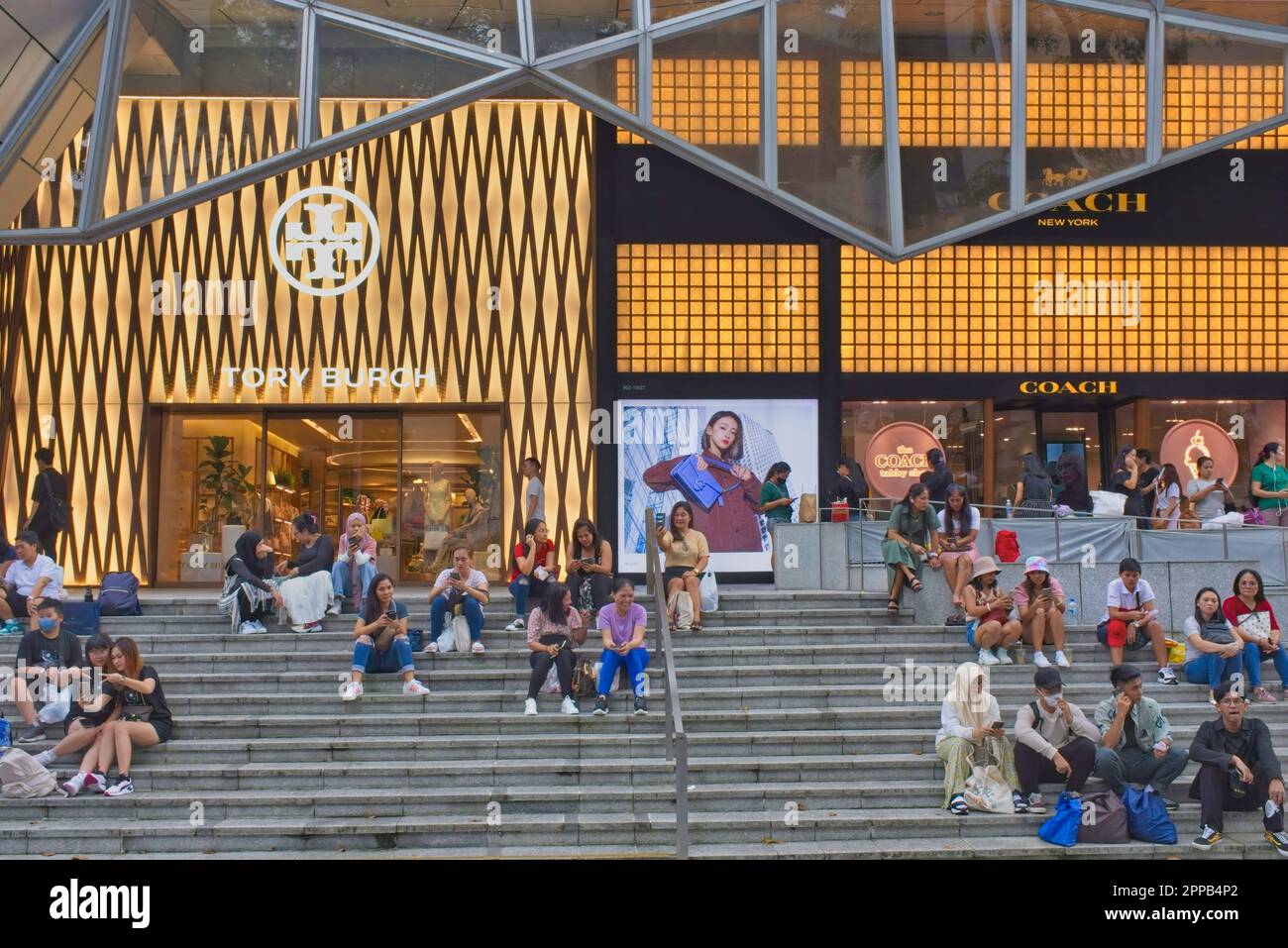 Gli amanti dello shopping si rilassano sui gradini di fronte a un outlet Tony Burch illuminato in Orchard Road, Singapore, la strada dello shopping più famosa della città Foto Stock