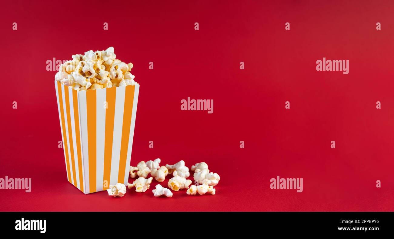 Secchio di cartone a strisce bianche gialle con gustoso popcorn al formaggio, isolato su sfondo rosso. Scatola con dispersione di grani di popcorn. Fast food, film, ci Foto Stock