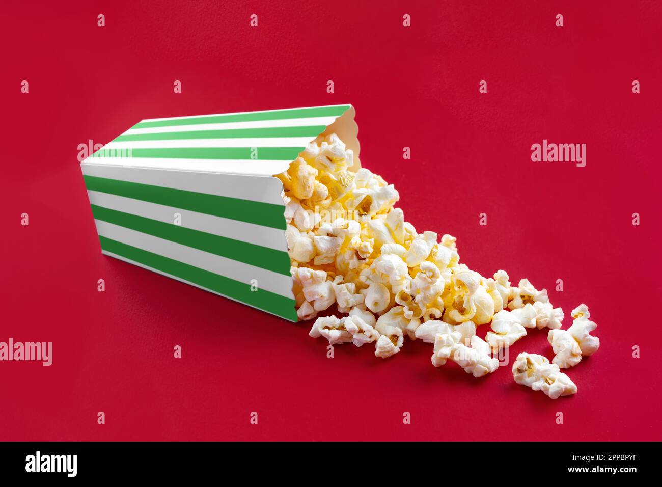 Gustoso popcorn al formaggio che cade da un secchio di cartone a strisce verdi, isolato su sfondo rosso. Dispersione di grani di popcorn. Film, cinema e intrattenimento Foto Stock