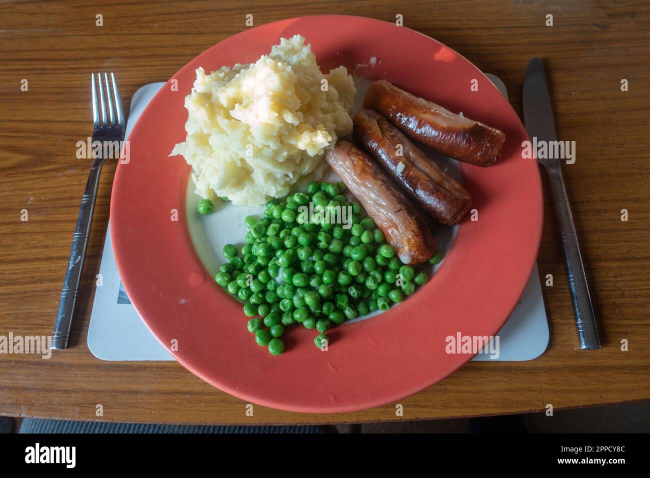 Salsicce, purè di patate e piselli su un piatto rosso, cibo confortevole per cena. Foto Stock