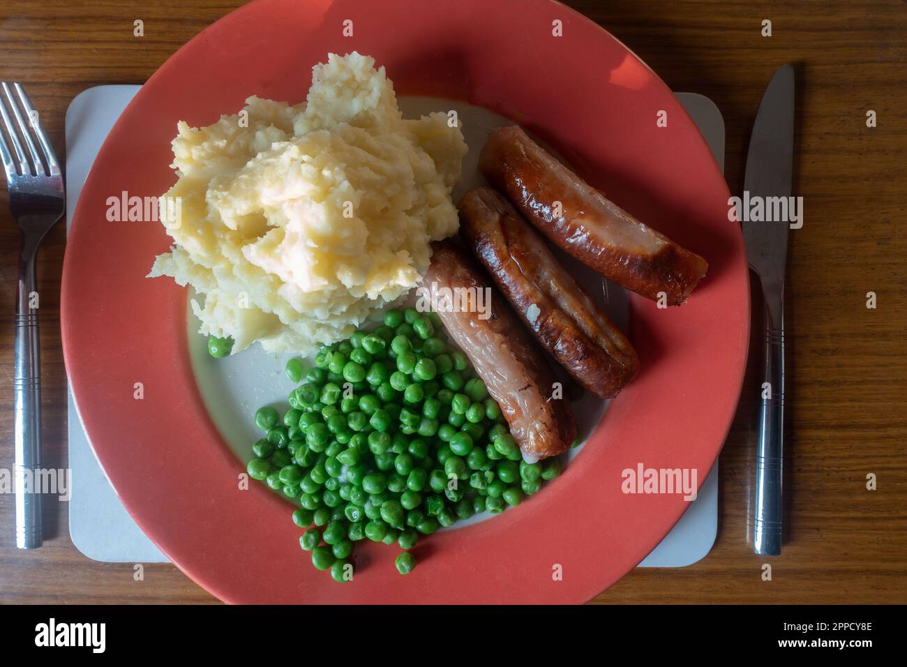 Salsicce, purè di patate e piselli su un piatto rosso, cibo confortevole per cena. Foto Stock