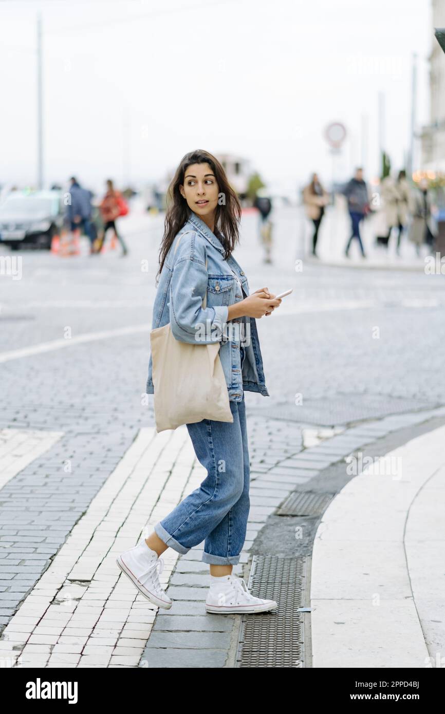 Giovane donna con borsa tote e smart phone che attraversa la strada Foto Stock