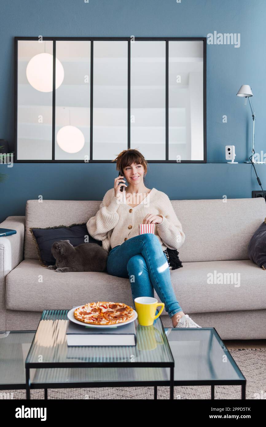 Donna sorridente che parla con uno smartphone seduto sul divano di casa Foto Stock
