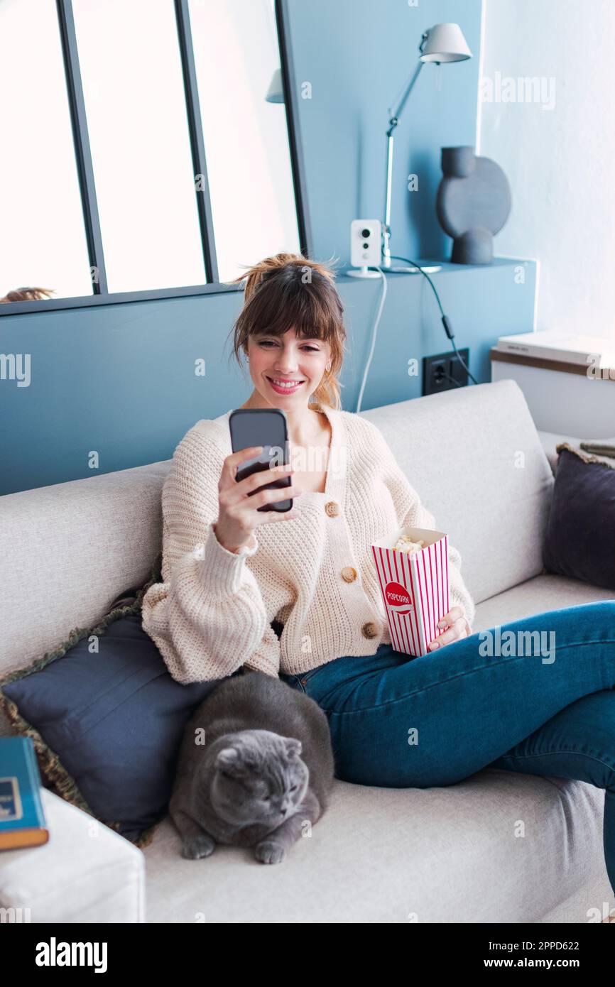 Donna sorridente con popcorn utilizzando smartphone seduto sul divano a casa Foto Stock