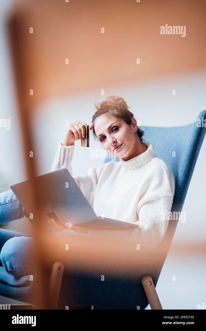 Freelance che tiene la carta di credito seduto con il computer portatile in poltrona presso l'ufficio di casa Foto Stock