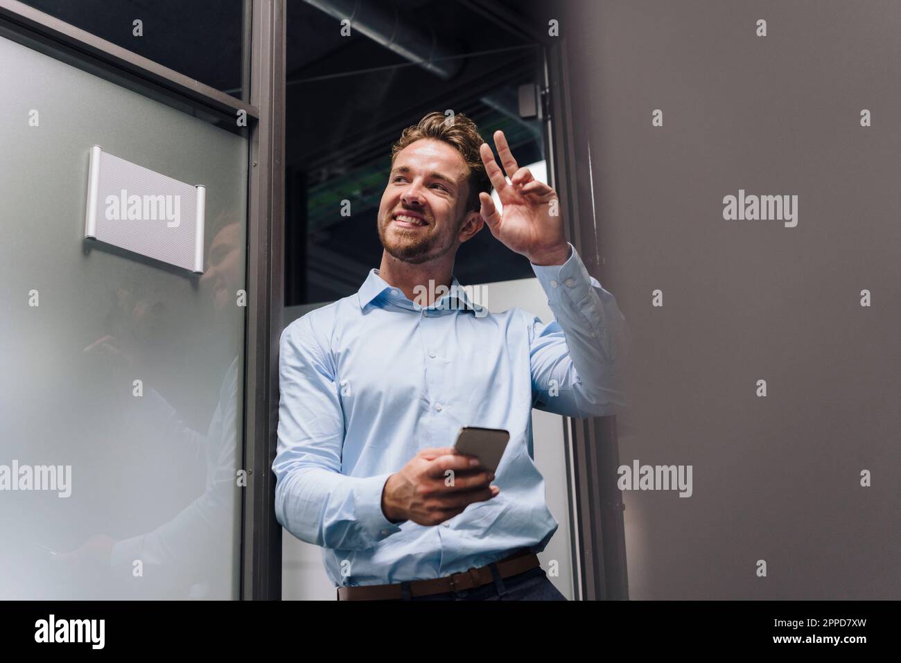 Uomo d'affari felice con smartphone gesturing da porta in ufficio Foto Stock