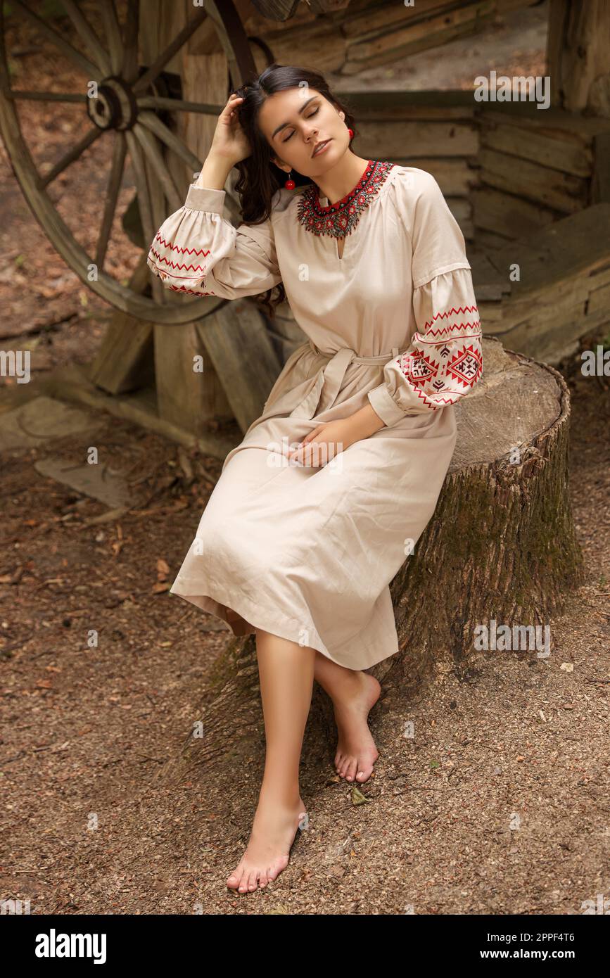 Bella donna con vestito ricamato seduta vicino al vecchio pozzo di legno in campagna. Vestiti nazionali ucraini Foto Stock