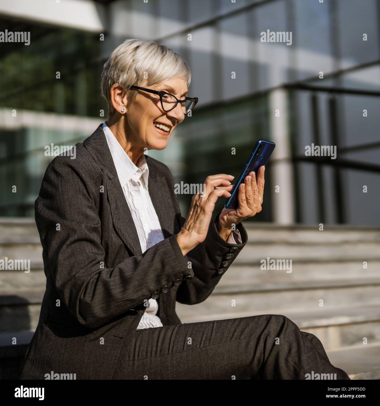 Donna d'affari matura seduta davanti all'edificio dell'azienda e alla messaggistica su smartphone. Foto Stock