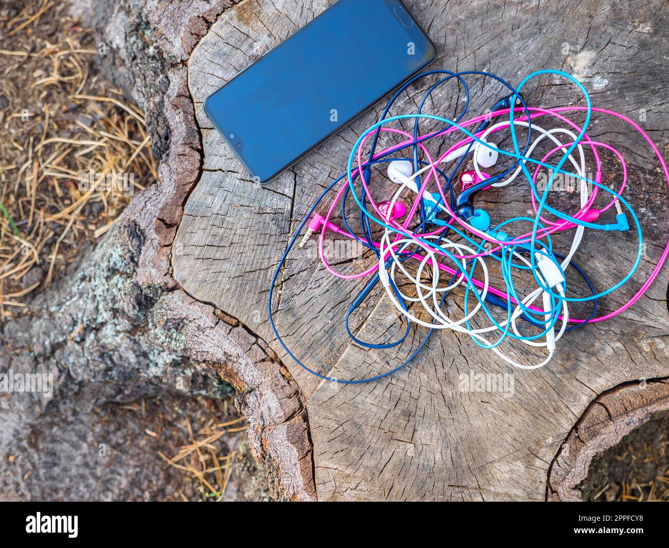 Un telefono e un nodo di quattro paia di auricolari rosa, blu, bianco e nero con fili intrecciati in un problema caotico e disordinato vicino a uno smartphone su un moncone. Foto Stock