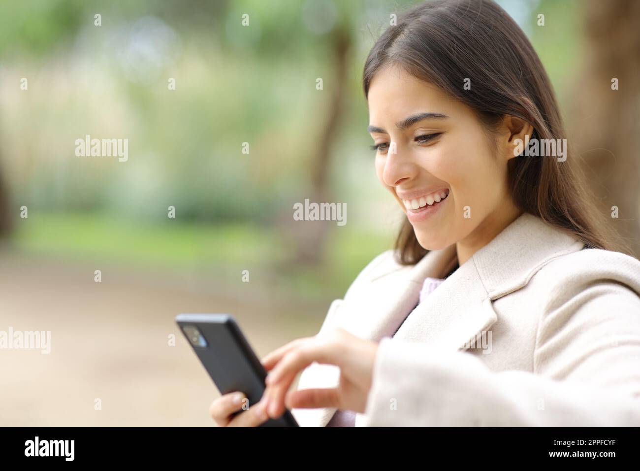 Donna felice in inverno utilizzando il telefono cellulare seduto in un parco Foto Stock