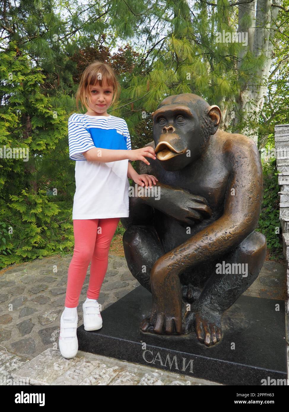 Belgrado, Serbia, 25 aprile 2021. Una bambina di 7 anni con i capelli biondi si trova accanto alla statua dello scimpanzé Sami, il simbolo dello zoo di Belgrado. Iscrizione - Sami, il soprannome della scimmia. Foto Stock