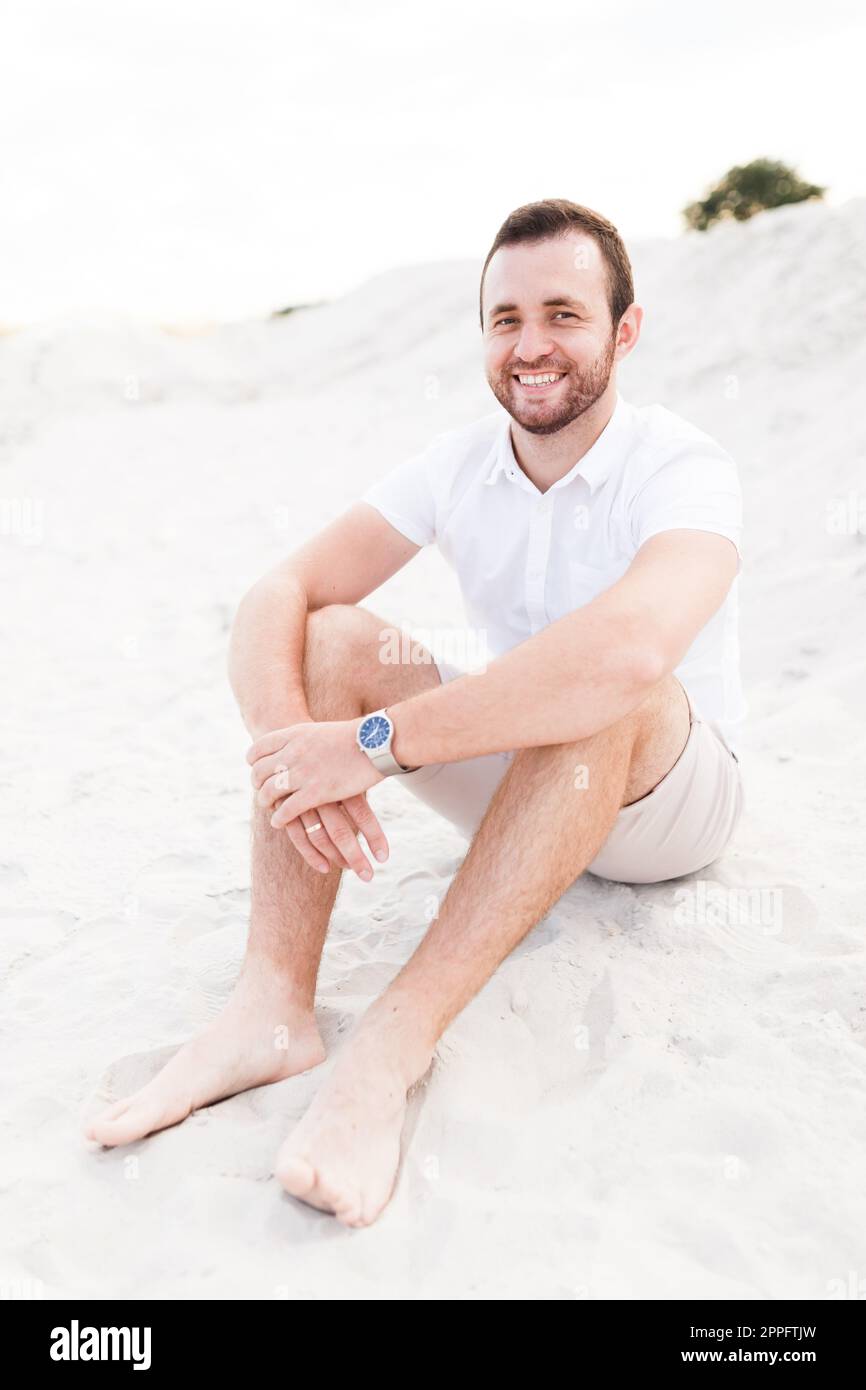 Giovane uomo felice beared in luce seduto sulla spiaggia Foto Stock