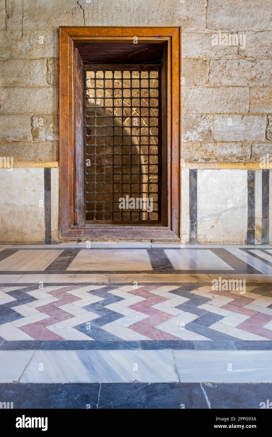 Finestra incassata in legno con griglia di ferro decorata su pareti in mattoni di pietra e pavimento decorativo colorato Foto Stock