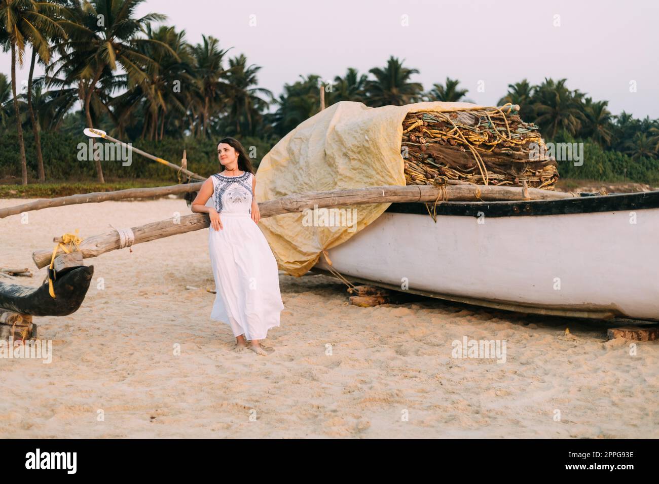Goa, India. Giovane donna caucasica in vestito bianco in piedi vicino alla barca da pesca e in posa per la macchina fotografica Foto Stock