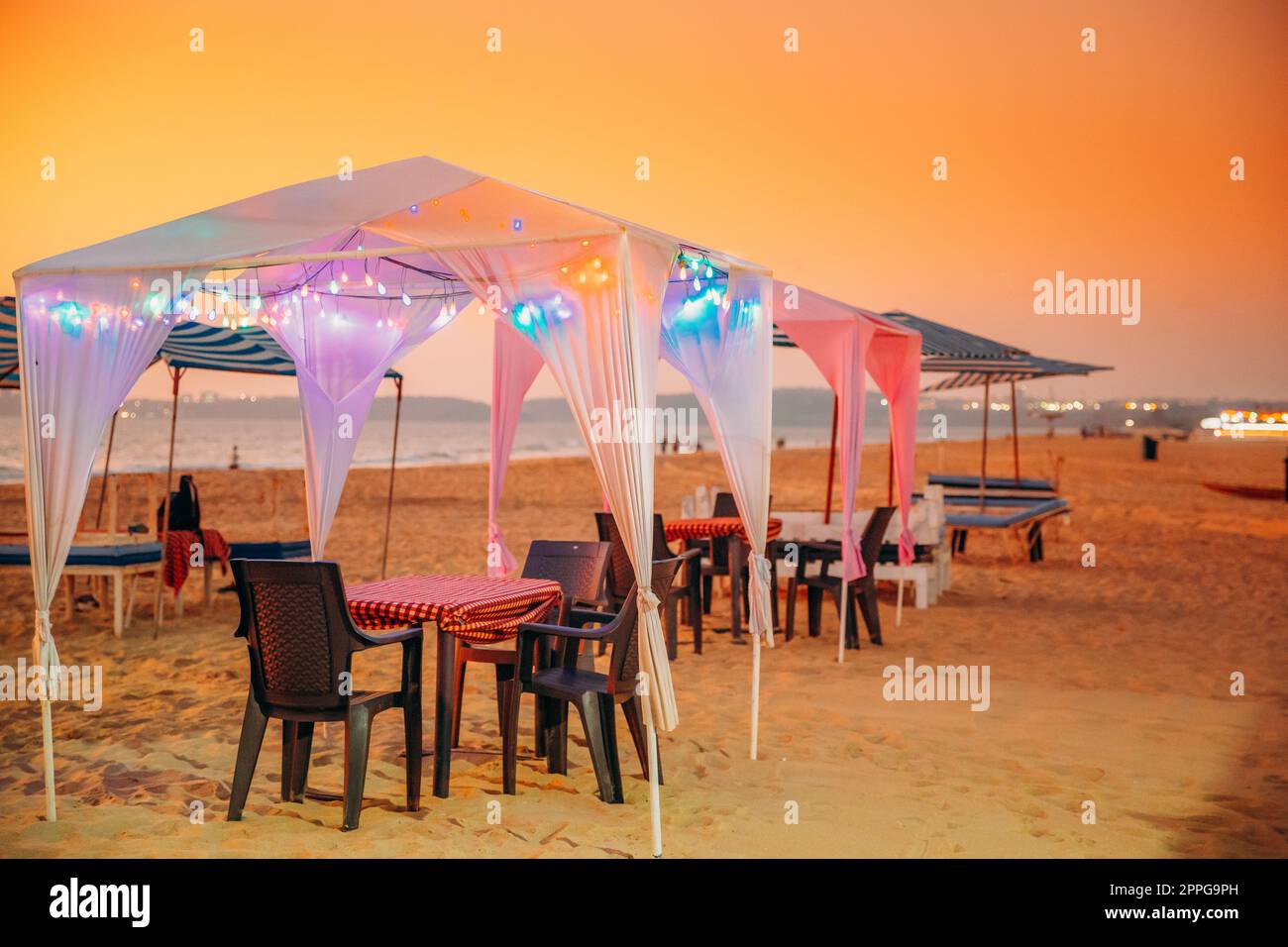 Goa, India. Tende da gazebo con lampada per i turisti sulla spiaggia con tavoli e sedie all'interno al tramonto Foto Stock