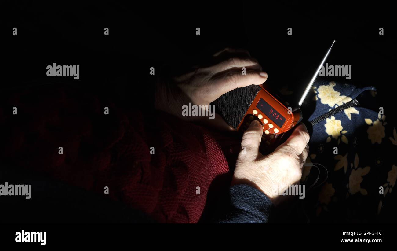 Ucraina: La nonna tiene la radio nelle mani seduta nel buio del blackout durante gli attacchi terroristici russi. Donna anziana in abiti invernali al buio sul divano con ricevitore radio a mano che ascolta le notizie Foto Stock