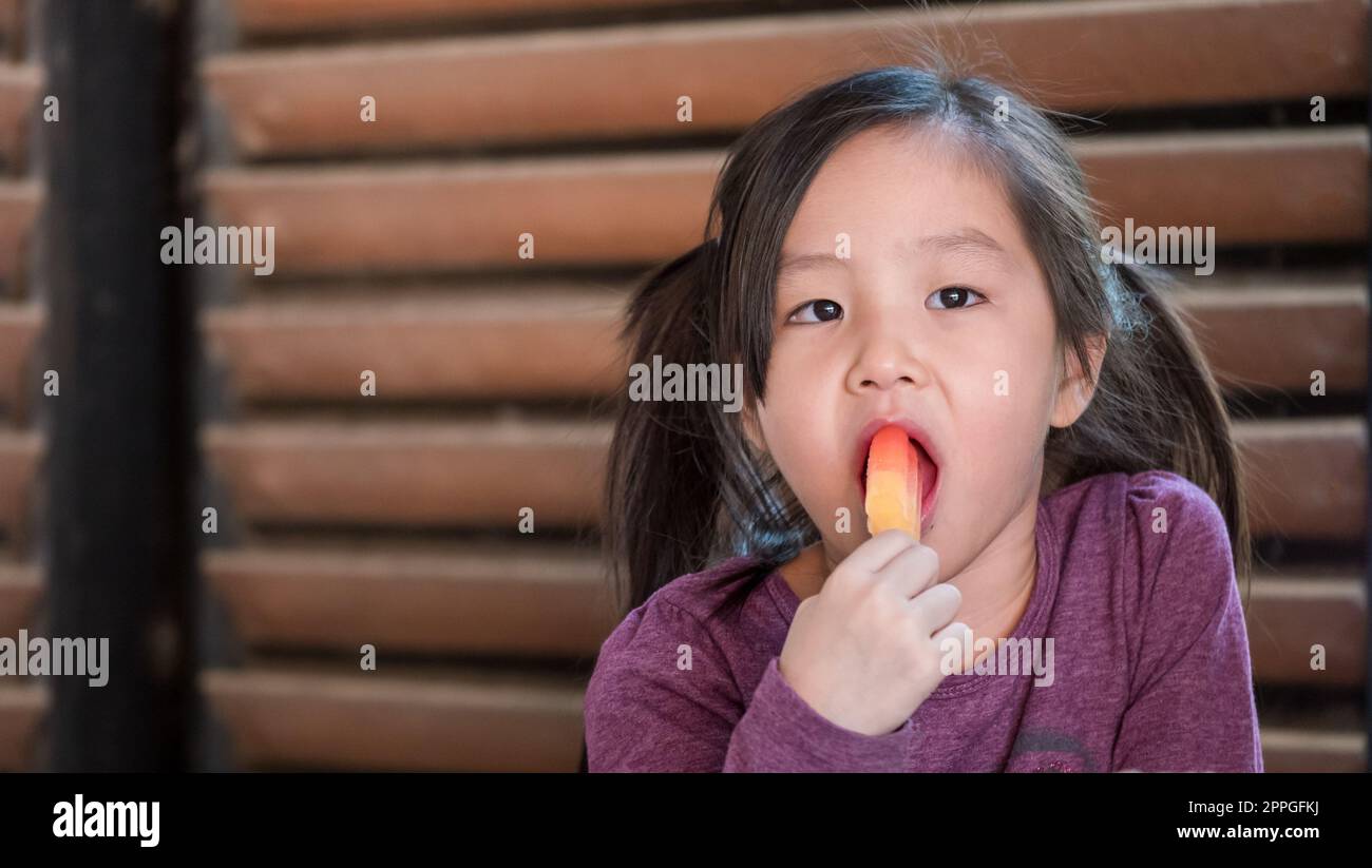 Piccola ragazza asiatica che mangia gelato, sfondo strisce tonalità legno Foto Stock