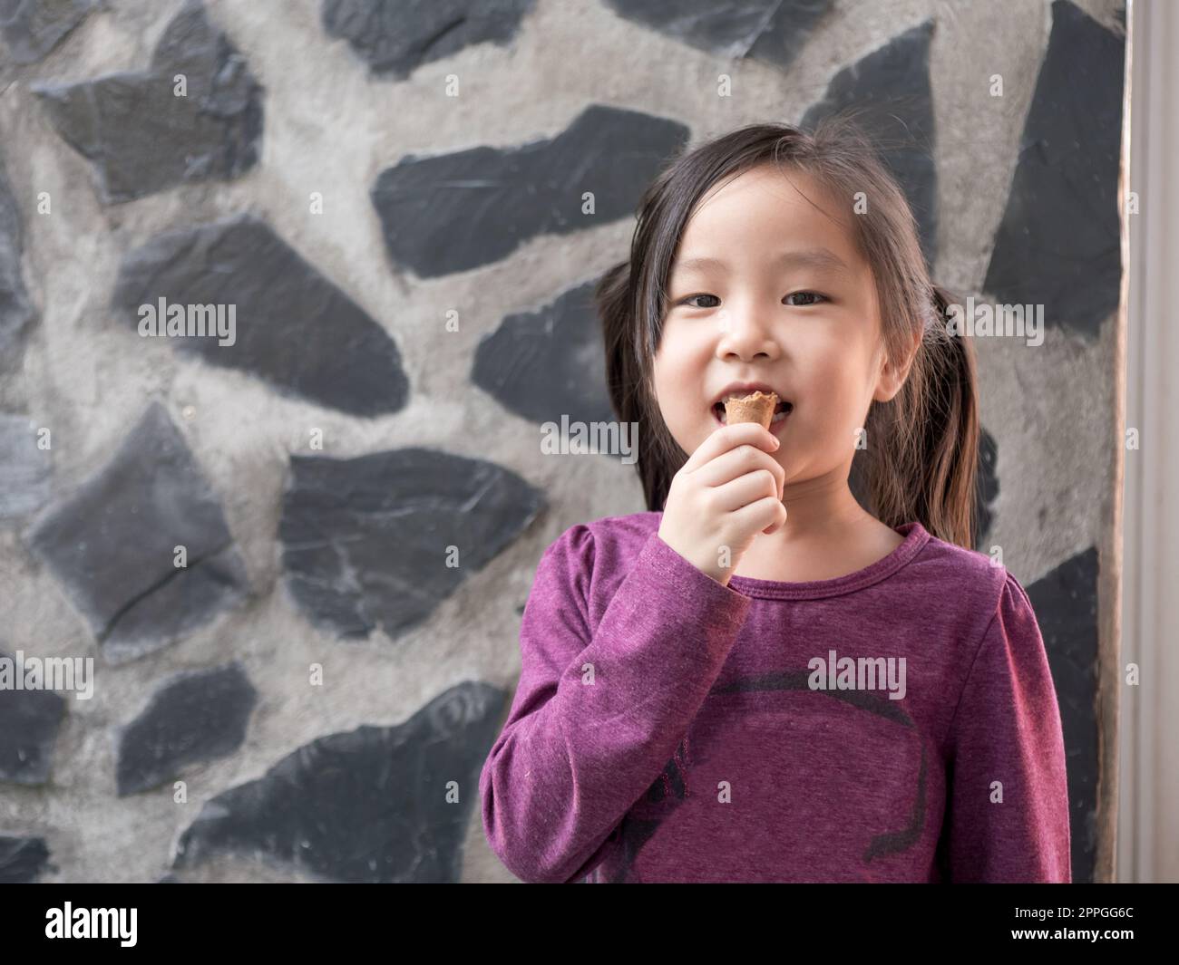 Piccola ragazza asiatica che mangia gelato, sfondo muro di pietra Foto Stock