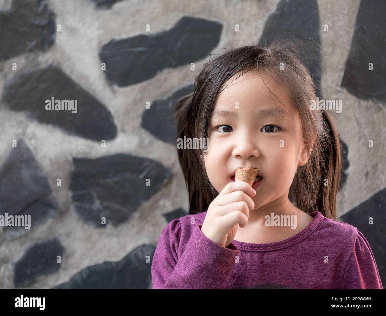 Piccola ragazza asiatica che mangia gelato, sfondo muro di pietra Foto Stock