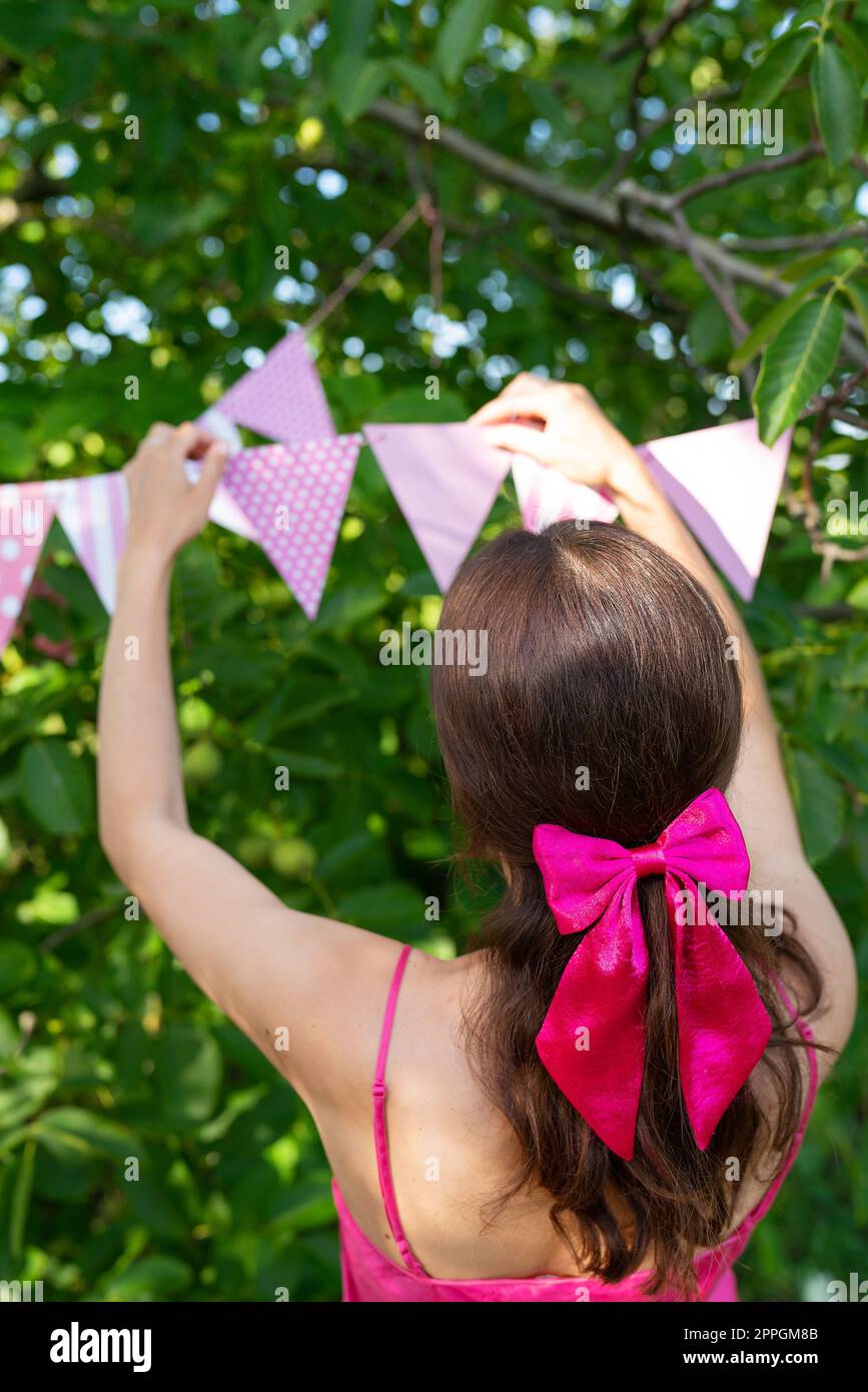 La ragazza appende bandiere triangolari di colore rosa sullo sfondo di un giardino verde. Una ragazza con un vestito rosa e un fiocco sui capelli. Decorazioni festive - concetto di celebrazione. Foto Stock