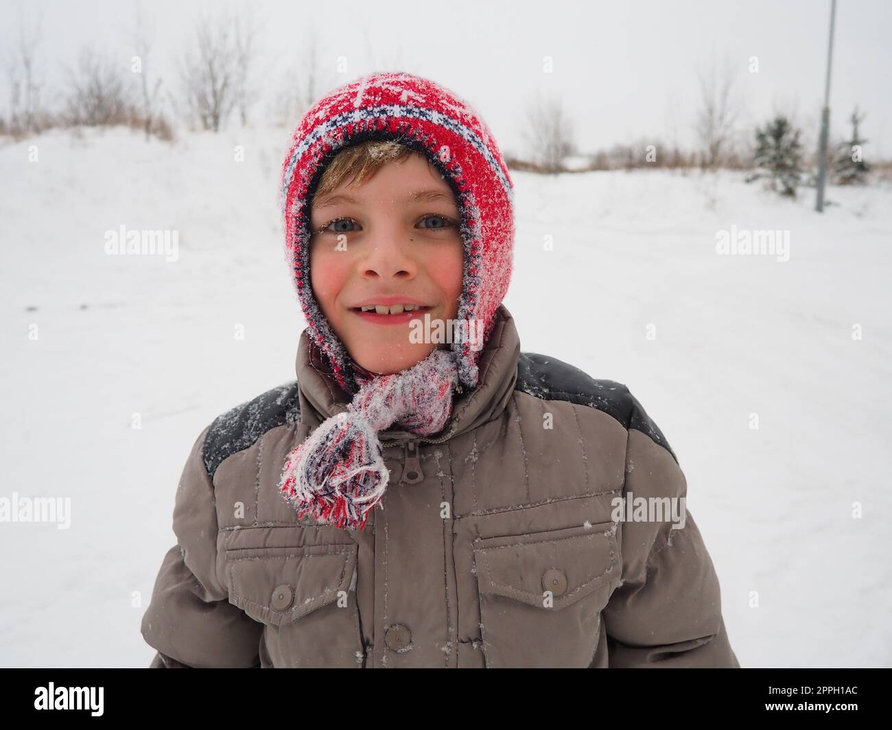 Un ragazzo carino di 8 anni con un cappello rosso a maglia con trecce e nappine. Il bambino sorride allegramente. Fiocchi di neve sul viso. Vacanze invernali, clima gelido e viaggi nel nord Foto Stock