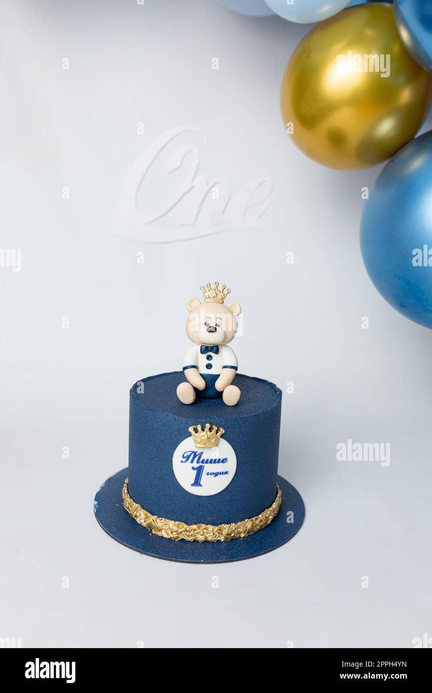 Una torta blu con un orsacchiotto sulla sommità con una corona su di essa. Iscrizione cirillica 'Misha ha ha 1 anni'. torta su sfondo bianco Foto Stock