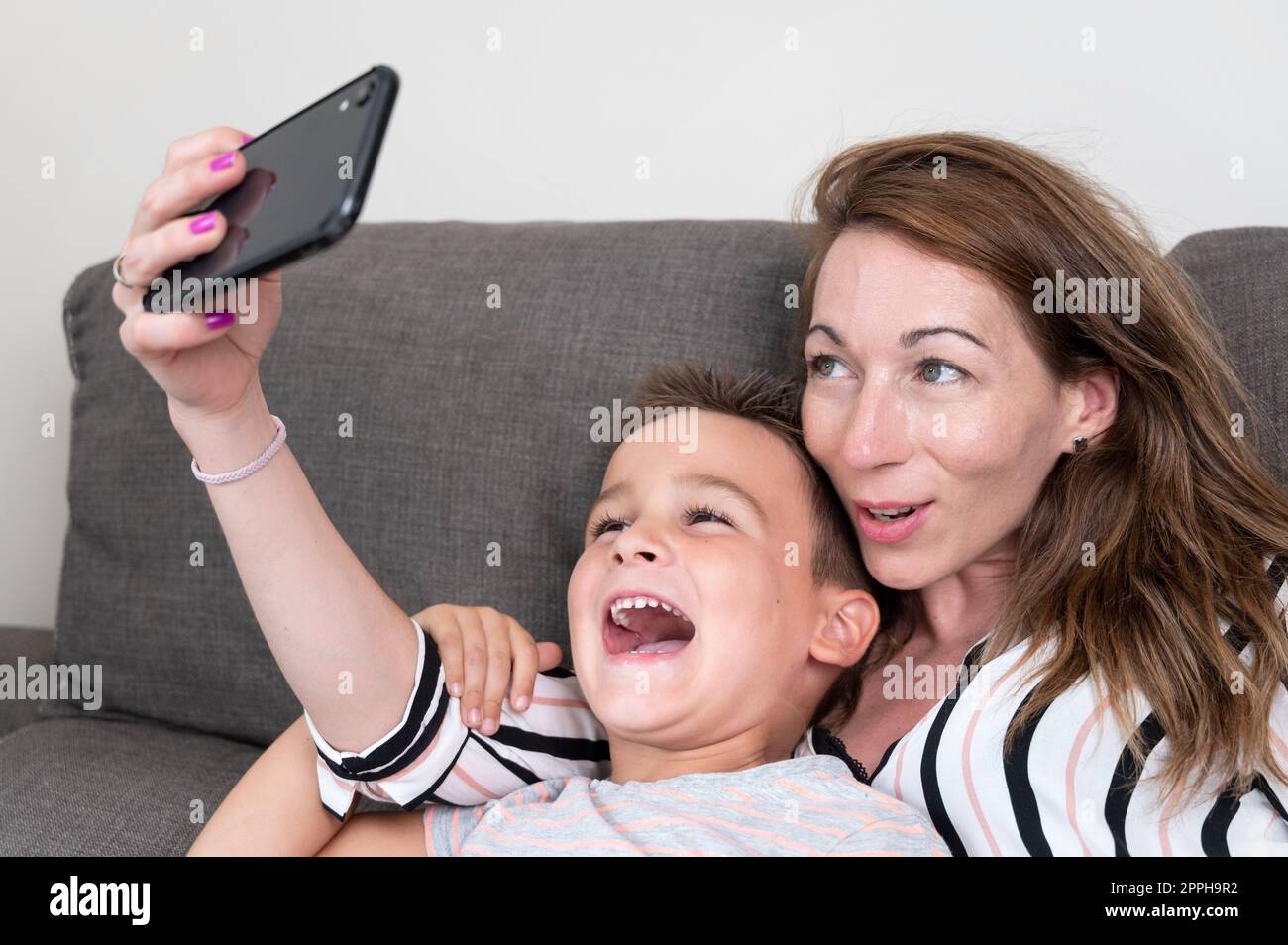 Una madre allegra con uno smartphone che fa selfie con il figlio piccolo che mostra il segno della vittoria. mamma e ragazzo si siedono sul divano usando gadget divertendosi a fare autoritratto passare del tempo insieme a casa. Foto Stock