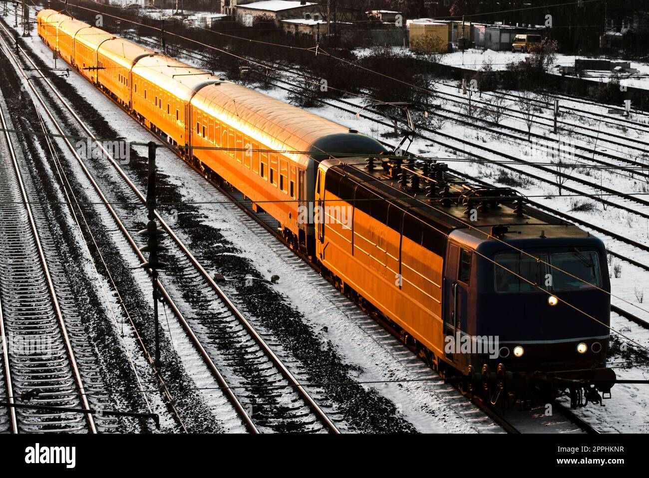 La locomotiva e le carrozze di un treno passeggeri viaggiano diagonalmente attraverso il quadro, illuminato di arancione dal tramonto o dal sole che sorge. Foto Stock