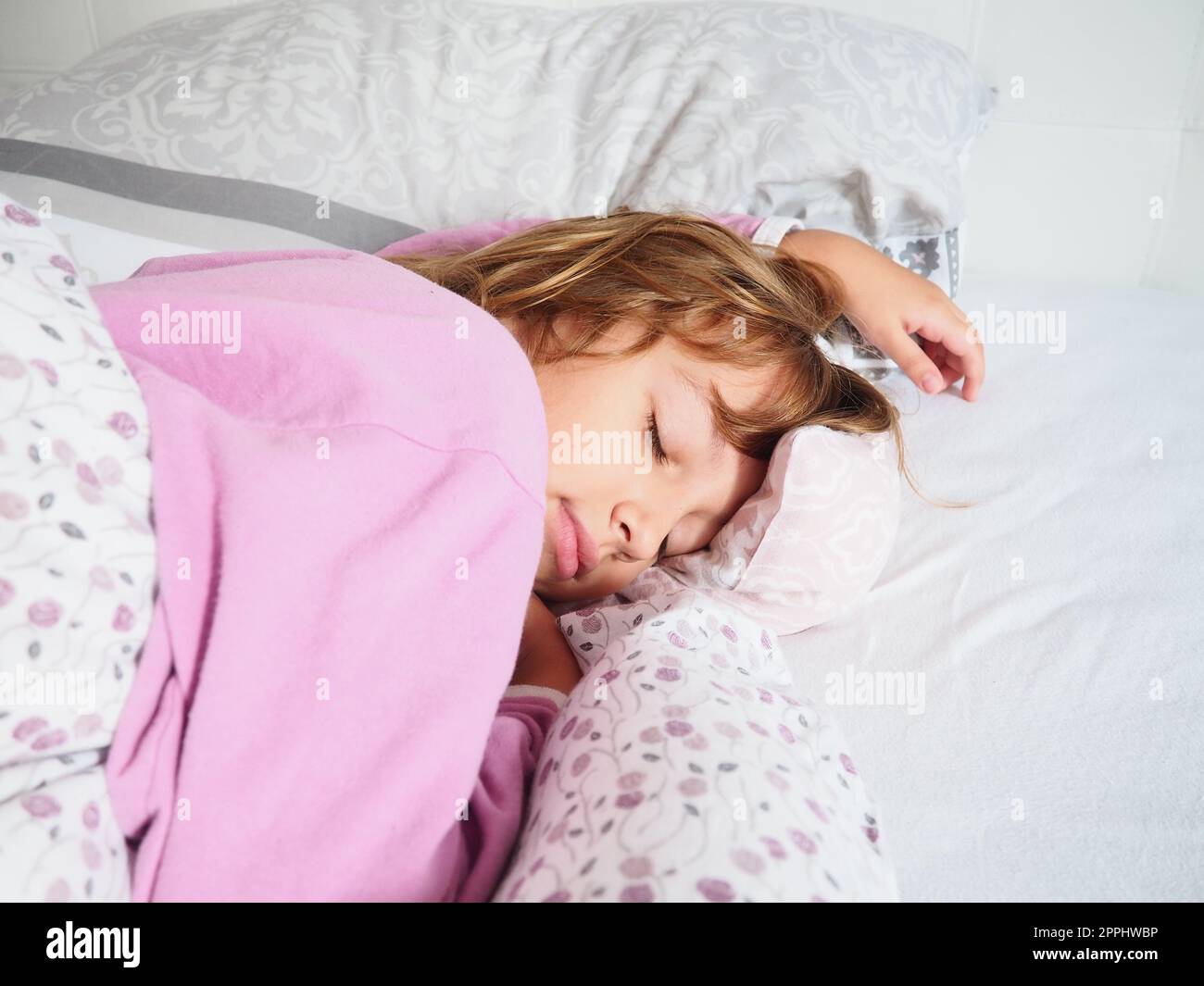 Una bella ragazza caucasica di 8 anni con capelli biondi, vestita con pigiama rosa, dorme su un letto con una soffice coperta, abbracciando un cuscino. La luce soffusa del sole del mattino scorre attraverso la finestra. Foto Stock