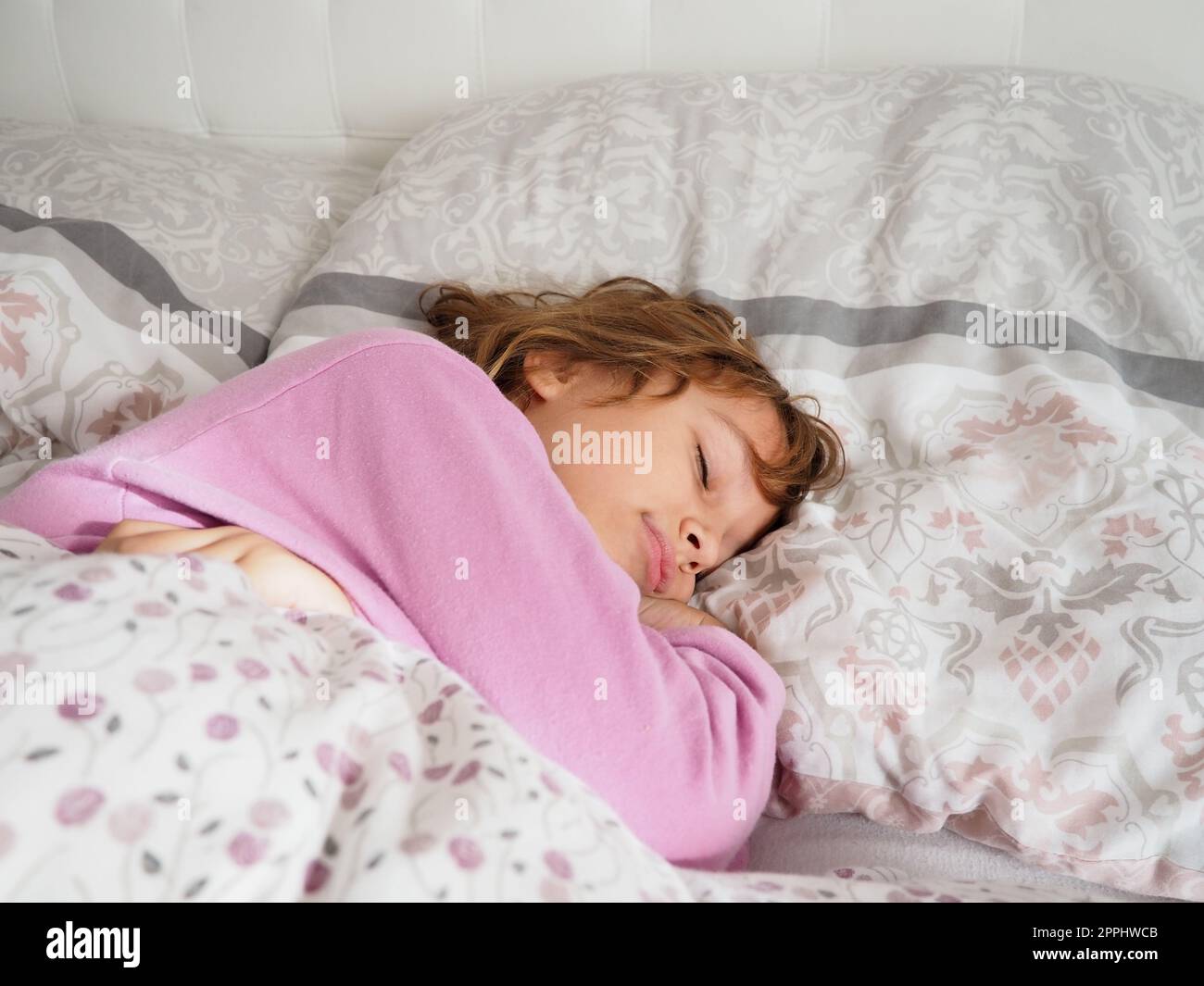 Una bella ragazza caucasica di 8 anni con capelli biondi, vestita con pigiama rosa, dorme su un letto con una soffice coperta, abbracciando un cuscino. La luce soffusa del sole del mattino scorre attraverso la finestra. Foto Stock