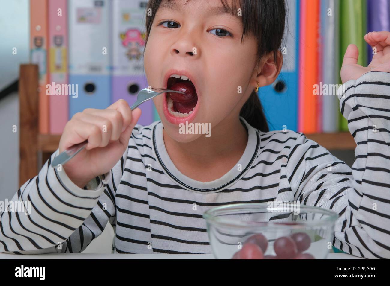 Bambina che mangia uva rossa fresca a casa nel soggiorno. Ragazza asiatica cute mangia frutta sana e latte per il suo pasto. Cibo sano nel bambino Foto Stock