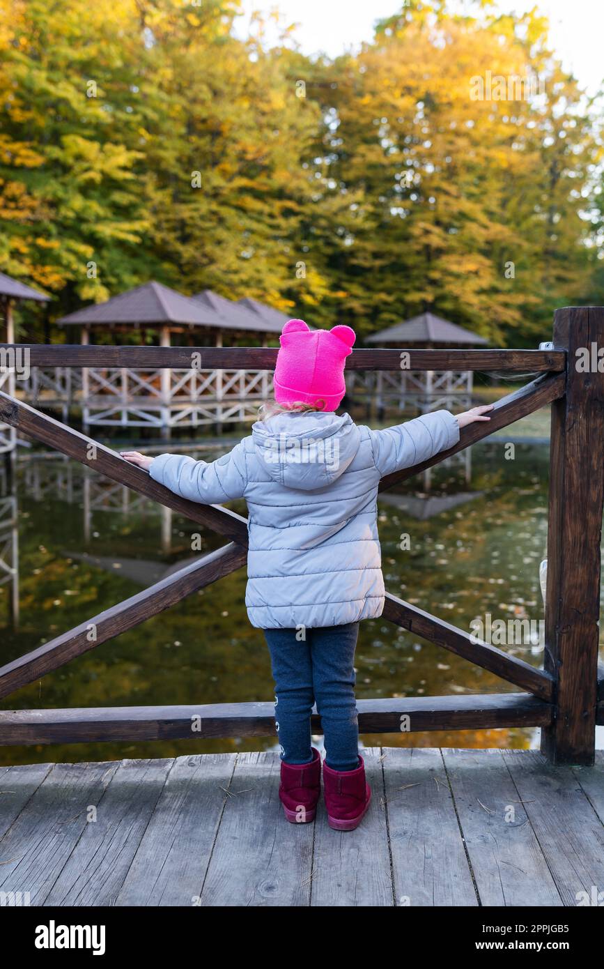 Una bambina con un cappello rosa si trova con la schiena vicino allo stagno e gode di una calda giornata autunnale. Foto Stock