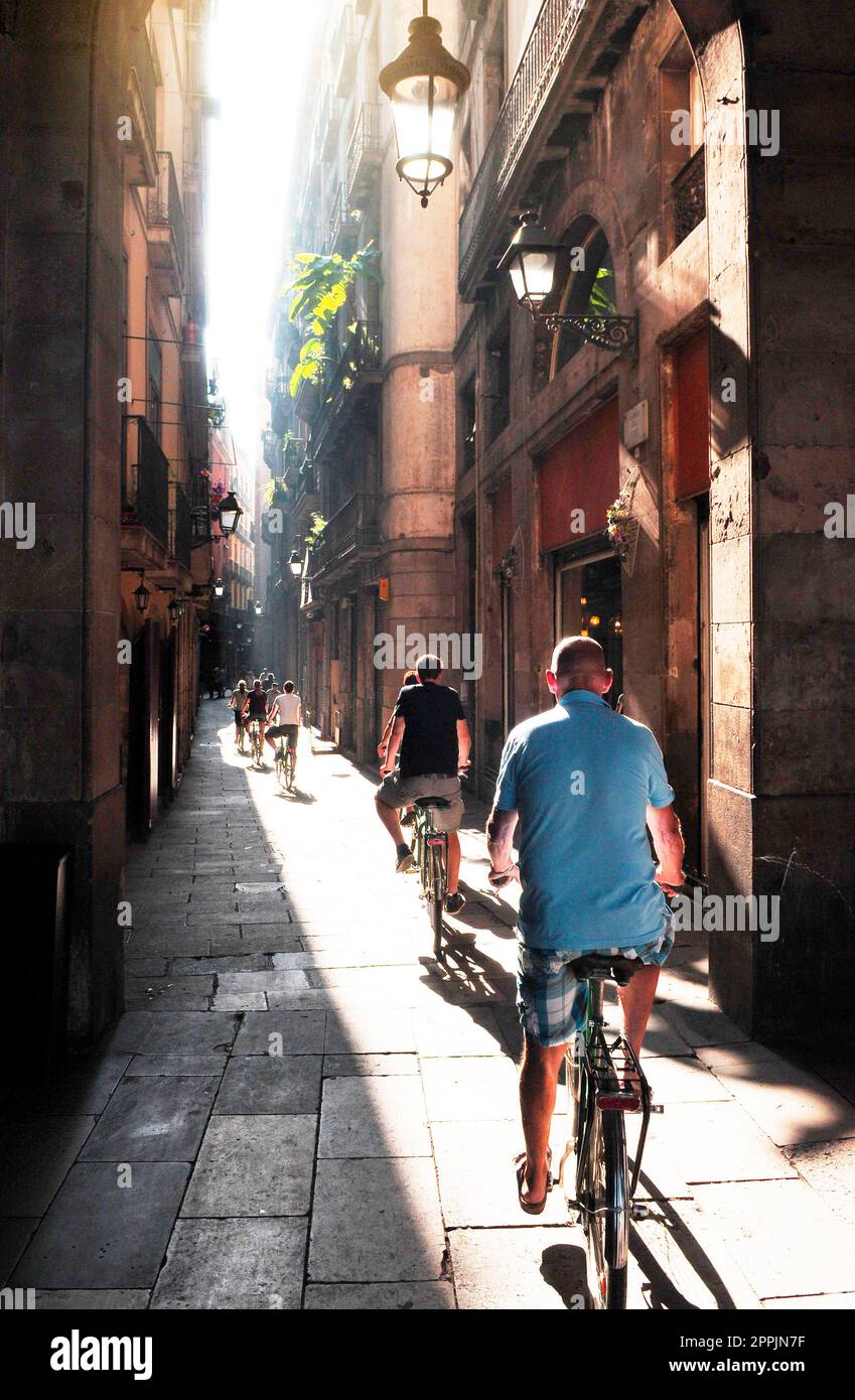 Gruppo di ciclisti sulla stretta strada di Barcellona, la luce del sole splende attraverso lo stretto spazio tra le case Foto Stock