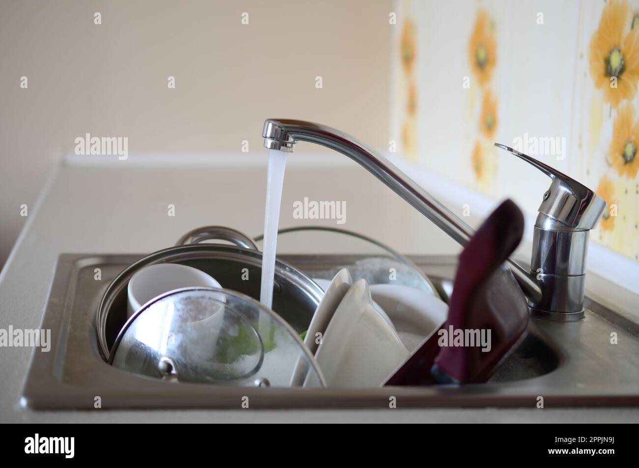 I piatti sporchi e gli elettrodomestici da cucina non lavati si trovano nell'acqua di schiuma sotto un rubinetto da un rubinetto della cucina Foto Stock