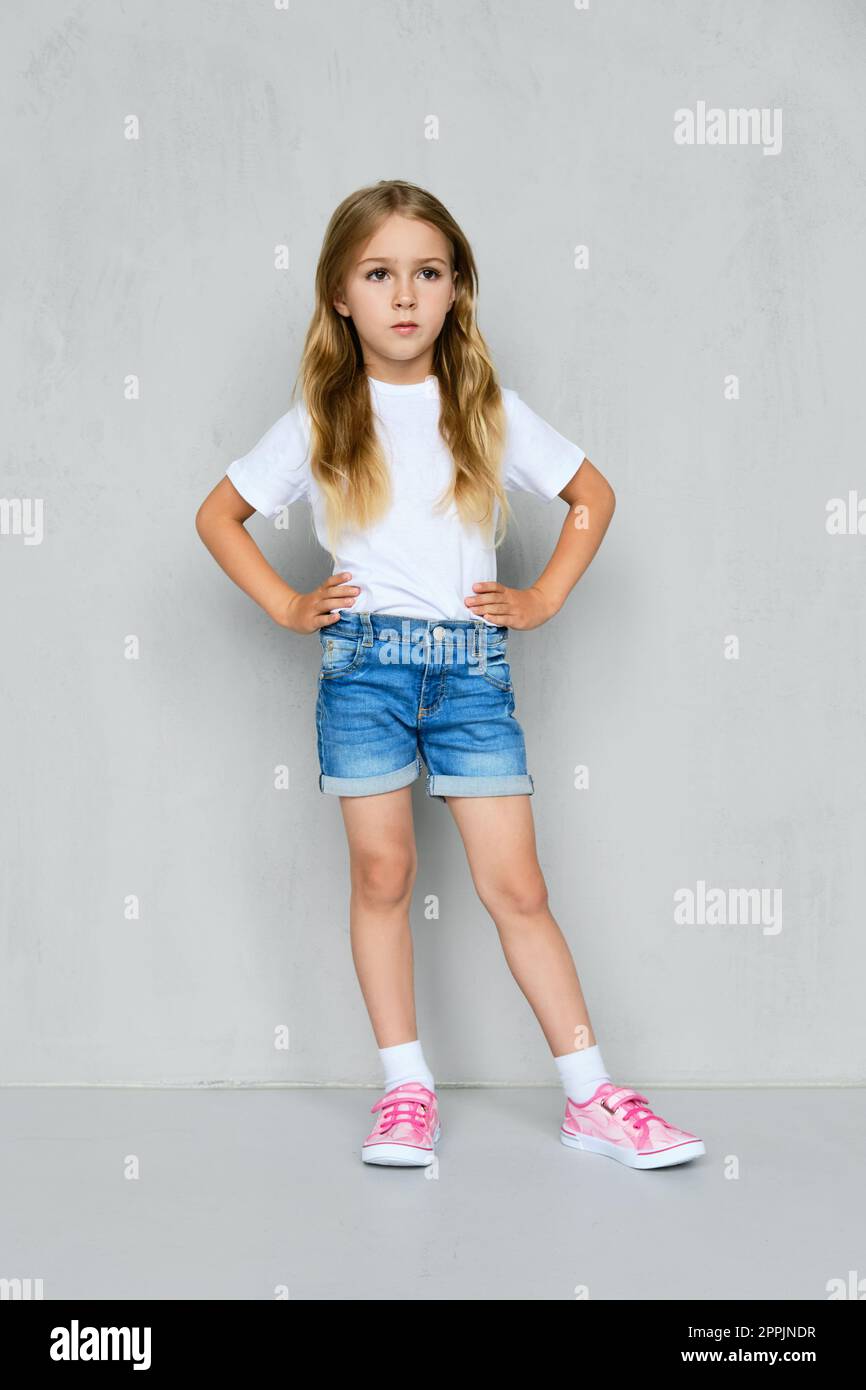 Bambina con t-shirt, pantaloncini jeans e sneakers in posa vicino al muro Foto Stock