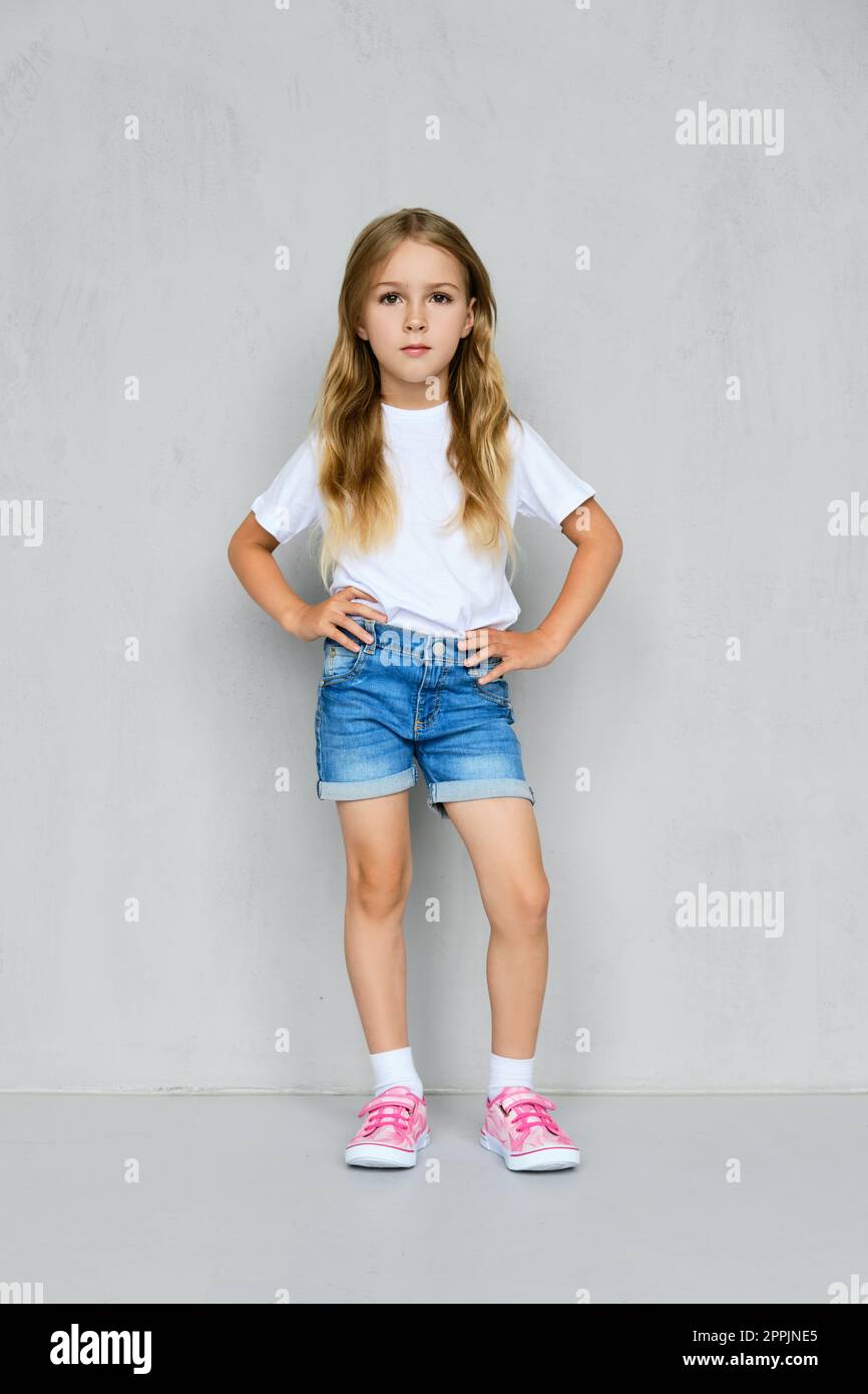 Bambina con t-shirt, pantaloncini jeans e sneakers in posa vicino al muro Foto Stock