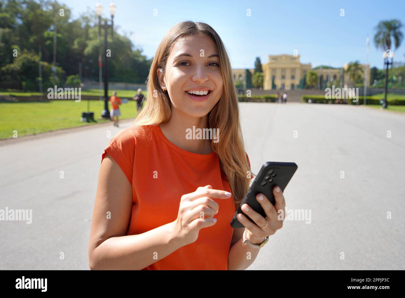 Felice bella giovane donna guardando la macchina fotografica e tenendo il telefono cellulare all'aperto con sfocato parco della città sullo sfondo. Tecnologia per lo stile di vita della città. Foto Stock