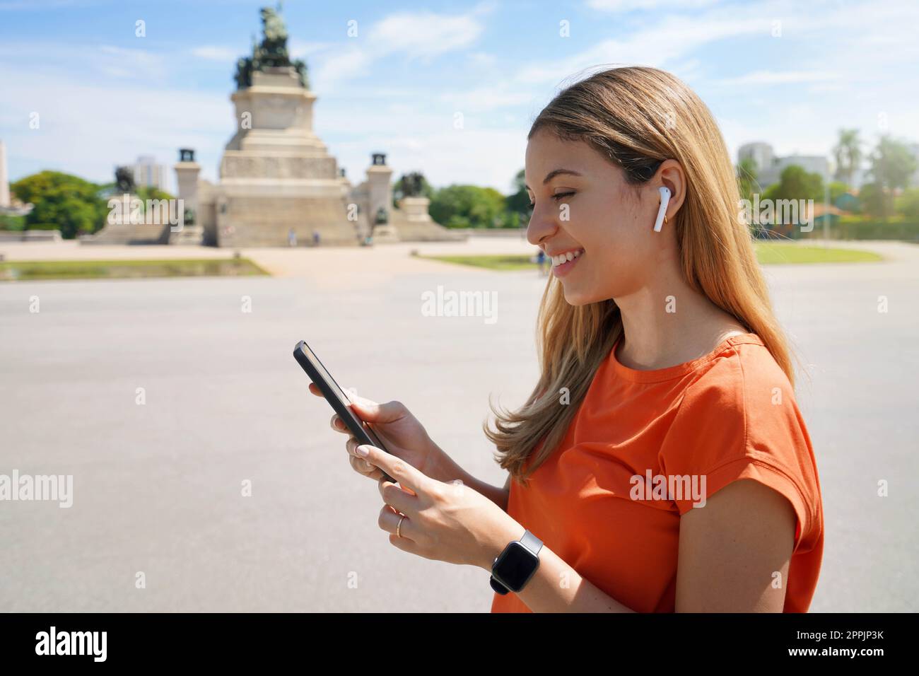 Vista laterale di una giovane donna ridente che sceglie la playlist guardando il suo smartphone nel parco cittadino di Sao Paulo, Brasile Foto Stock