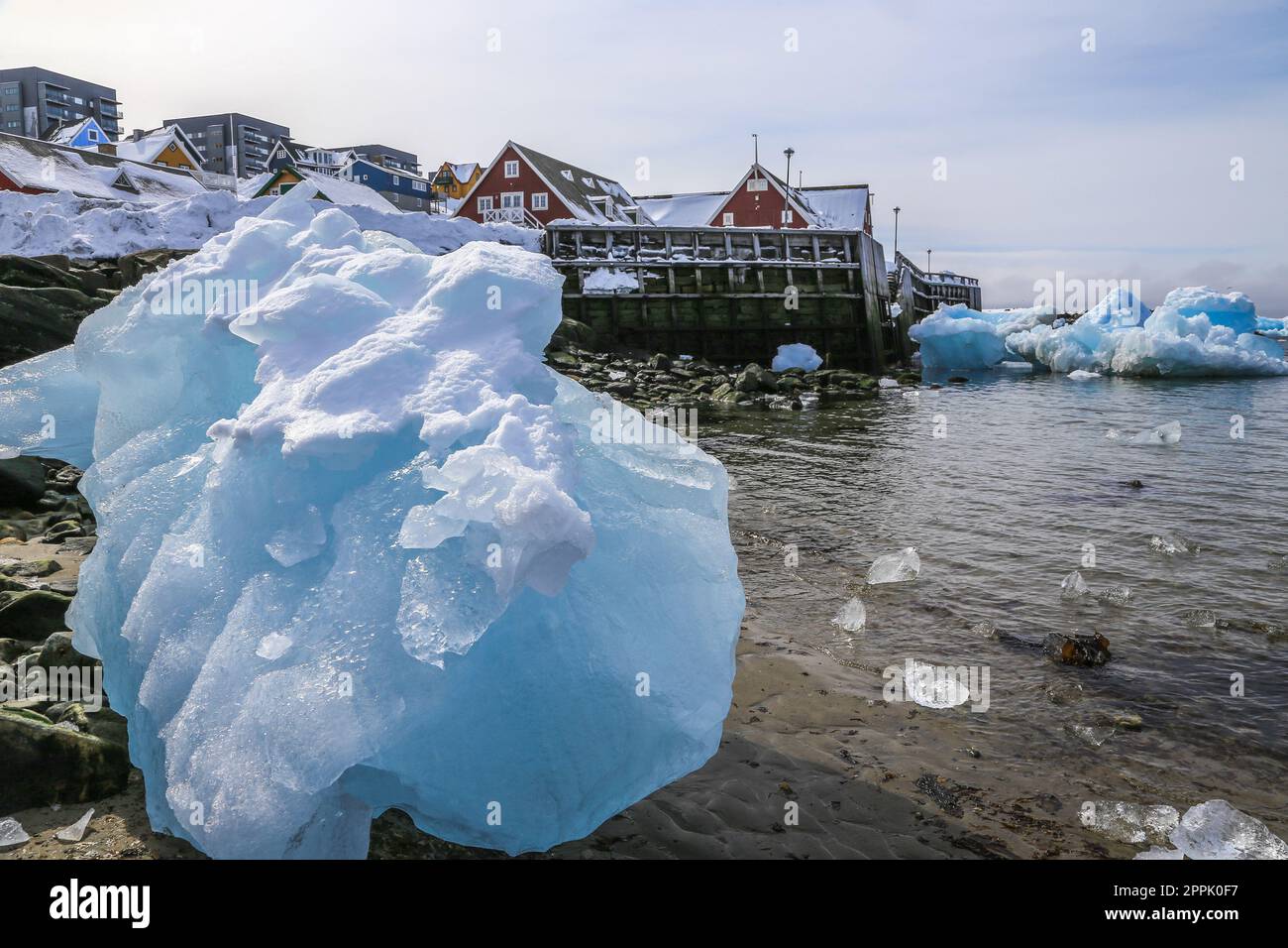 Grande pezzo di ghiaccio blu che stabilisce tra i sassi sulla riva con Inuit moderno edificio sulla collina al fiordo, città di Nuuk, Groenlandia Foto Stock