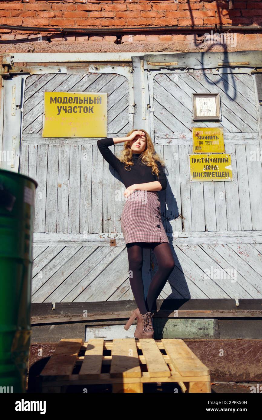 Una ragazza attraente si trova vicino alla porta della fabbrica con targhette di avvertenza e copre gli occhi con la mano. Foto Stock