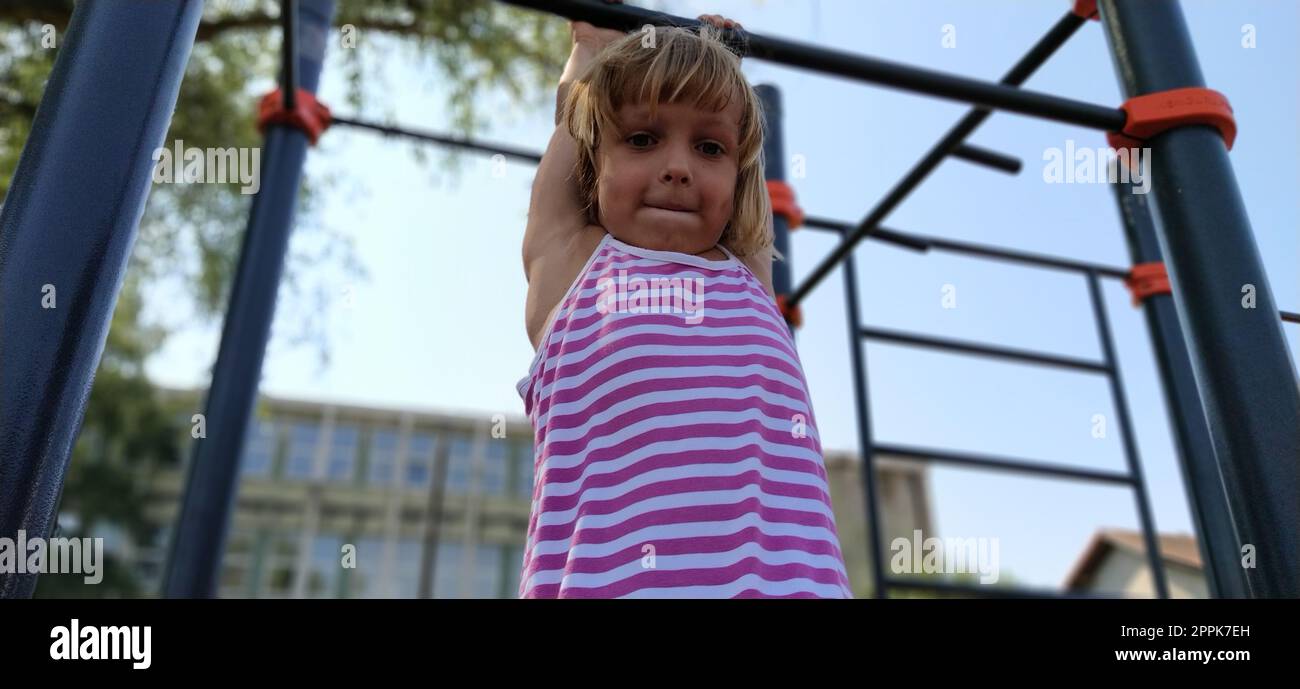 Un bambino con un vestito a righe pende su una barra di metallo e cerca di tirare le braccia. Sport o parco giochi. Palestra all'aperto Foto Stock