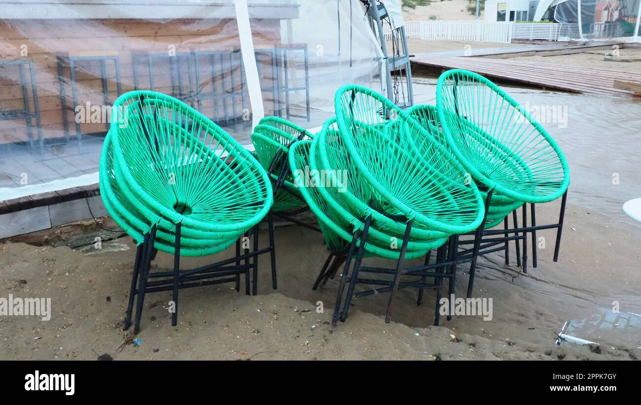 sedie in plastica verde vimini con gambe nere in un bar su una spiaggia sabbiosa dopo una tempesta. I mobili sono impilati l'uno sull'altro in quanto il caffè sulla spiaggia è chiuso. Fine della stagione balneare in hotel. Foto Stock