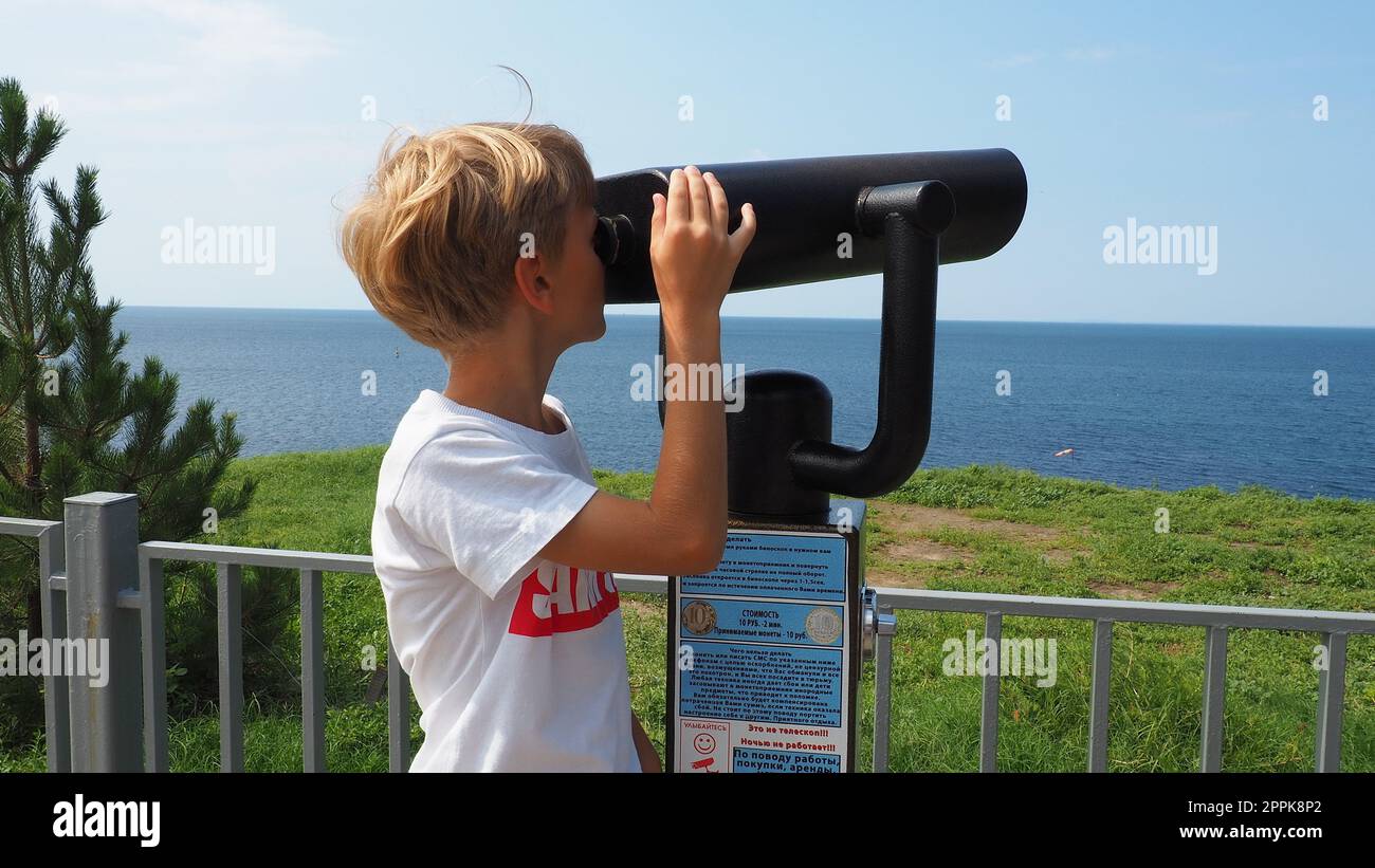 Anapa, Russia, 23 agosto 2021 Un ragazzo guarda attraverso un telescopio all'aperto. Un bambino caucasico di 9 anni tiene un grande telescopio marino con le mani. La strada del villaggio turistico e i turisti che camminano Foto Stock