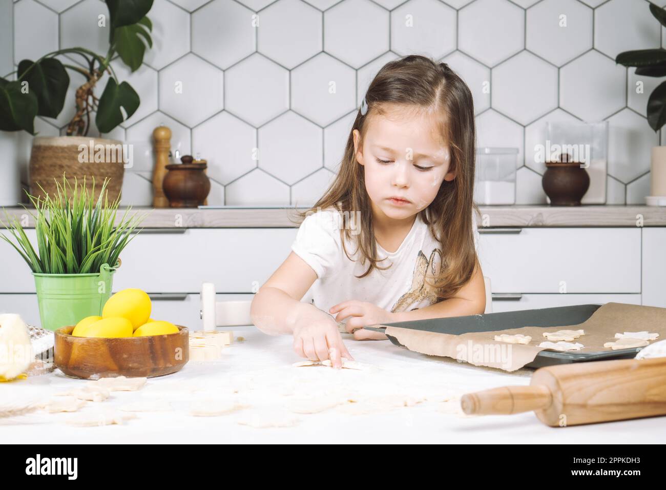 Ritratto di una bambina studiosamente piccola, cucina con sedia a sdraio, preparazione di diversi biscotti di pasta a forma di pasqua, padella Foto Stock