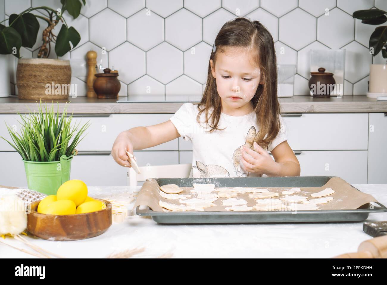 Ritratto di una bambina studiosamente piccola, cucina con sedia a sdraio, preparazione di diversi biscotti di pasta a forma di pasqua, padella Foto Stock