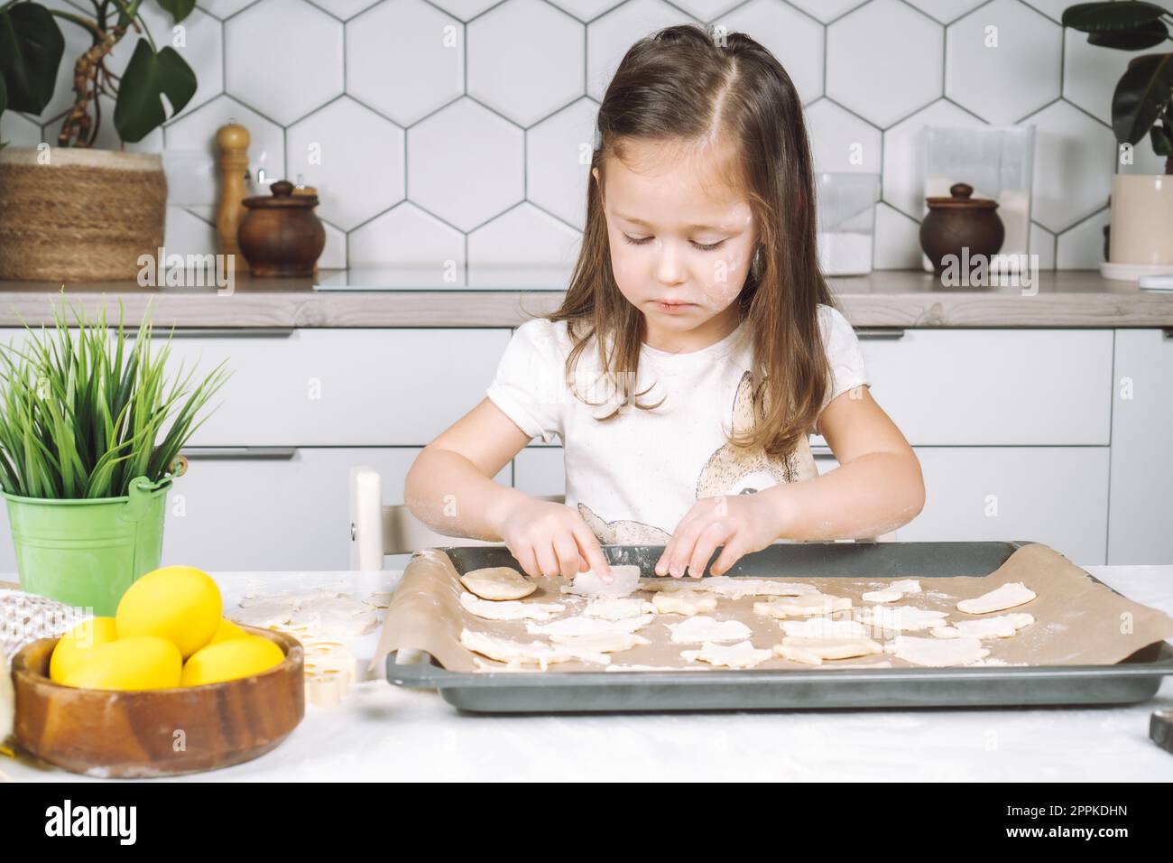 Ritratto di una bambina studiosamente piccola, cucina con sedia a sdraio, preparazione di diversi biscotti di pasta a forma di pasqua, padella Foto Stock