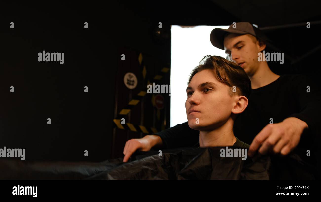 Uomo caucasico in camicia seduto sulla sedia mentre il barbiere chiude il suo cliente con peignoir nero nel salone. Servizio di parrucchiere in un moderno barbiere con illuminazione buia e vista laterale calda e luminosa Foto Stock