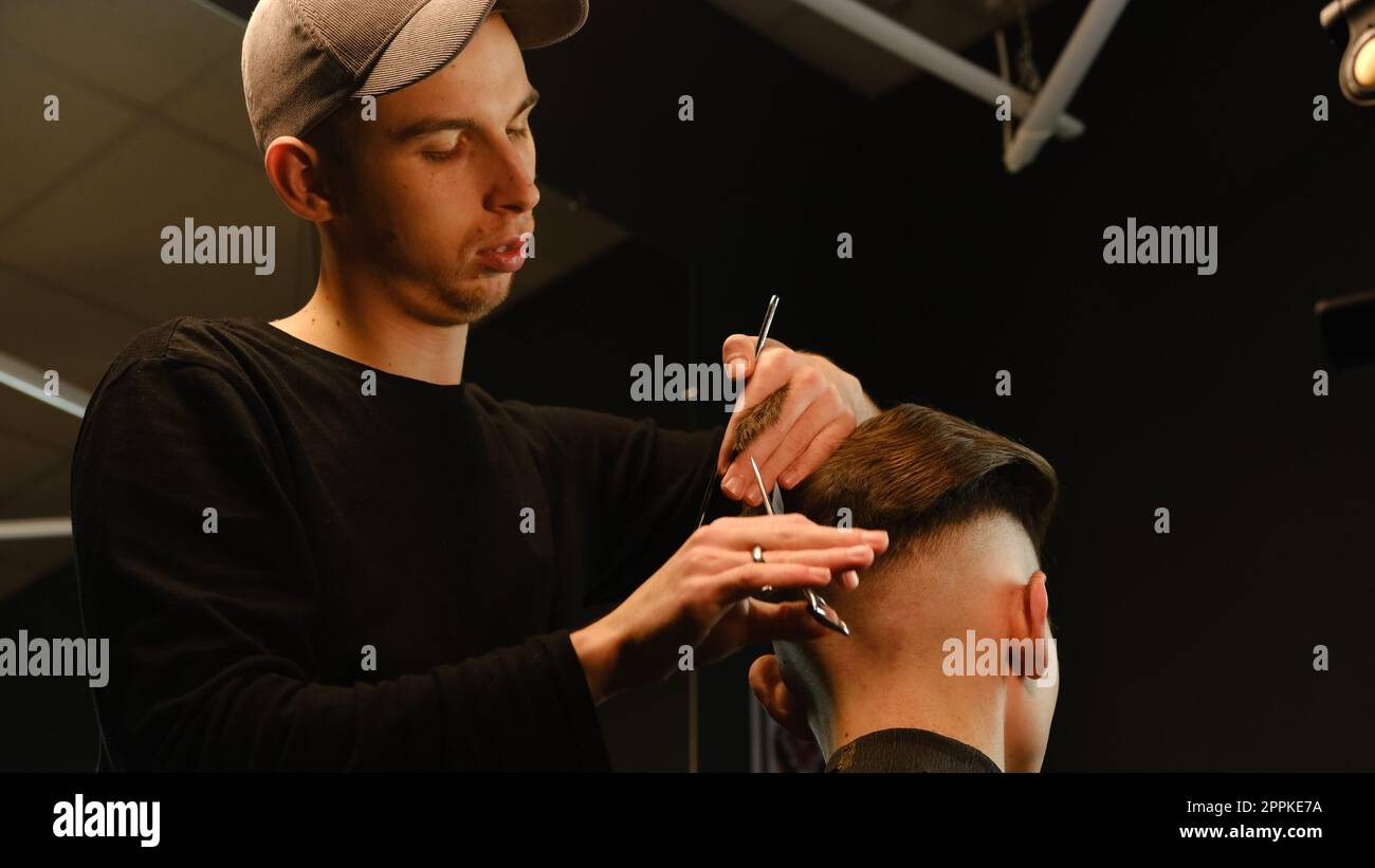 Il barbiere parrucchiere si occupa dell'acconciatura e dell'acconciatura con forbici e pettine. Concept Barbershop. vista posteriore con illuminazione a tasti scuri e luce calda Foto Stock