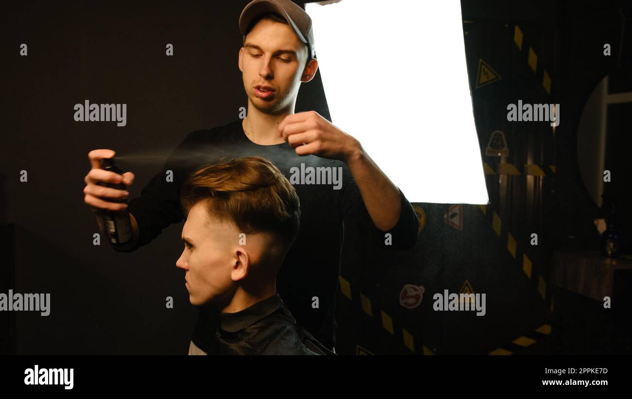 Il barbiere prepara lo styling dei capelli con lo spray dopo il taglio al barbiere. Giovane bell'uomo caucasico che ottiene un taglio di capelli in un moderno parrucchiere da parrucchiere professionale Foto Stock