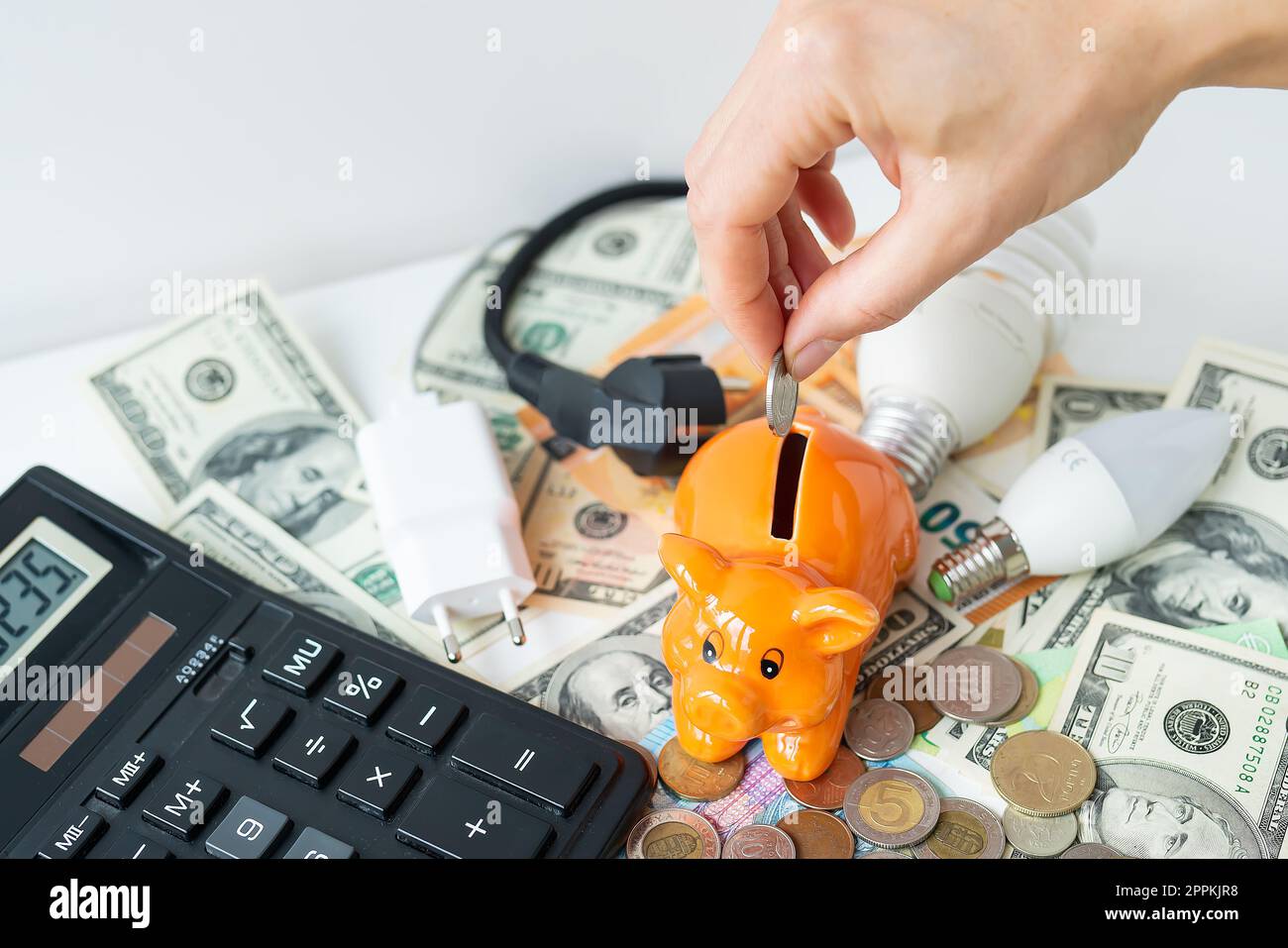 La mano di una donna mette una moneta in un salvadanaio arancione sotto forma di maiale, risparmiando denaro. Calcolatrice, banconote in euro e in dollari, monete. Tempo fiscale, pagamento fattura, calcolatore per il conteggio. Foto Stock
